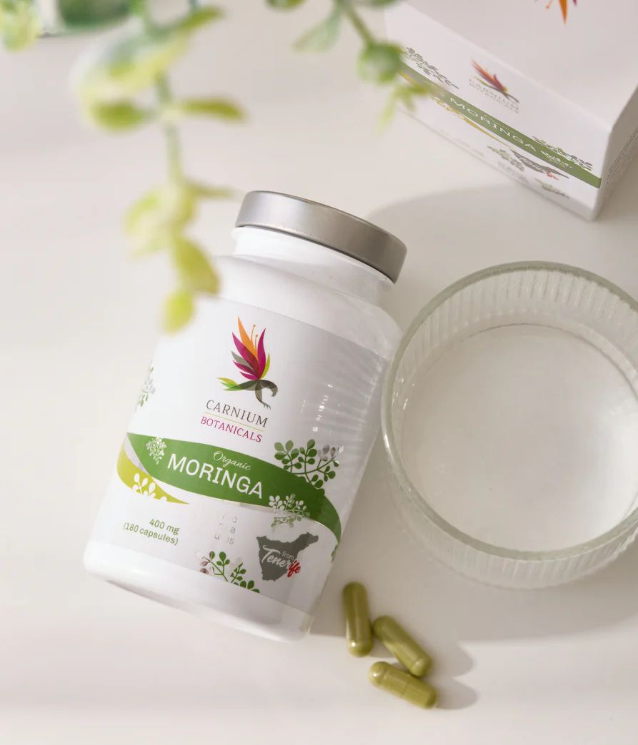 Weiße Flasche mit Kapseln, daneben Kapseln und ein Glas Wasser. Aufschrift: Carnium Botanicals, Moringa.