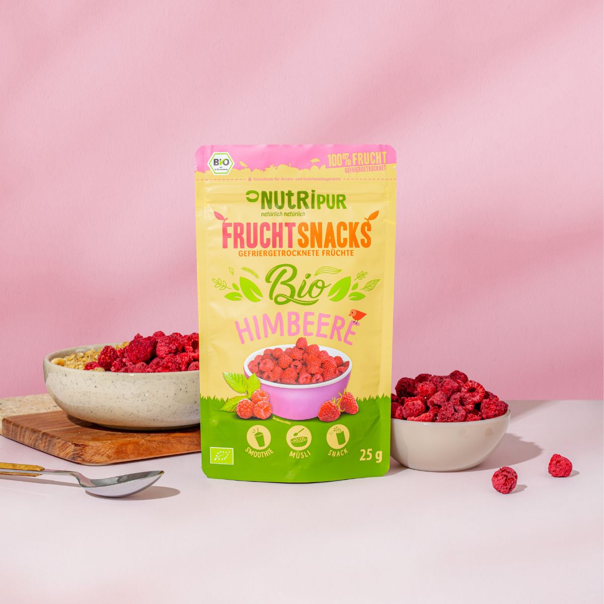 Beutel mit gefriergetrockneten Bio-Himbeeren, Schale mit Himbeeren, Schale mit Müsli und Himbeeren, Holzteller mit Löffel.