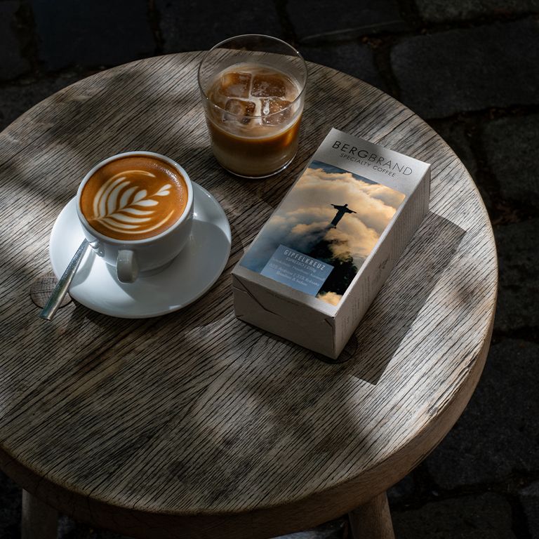 Kaffee und Getränk auf Tisch. Packung BERGBRAND Gipfelkreuz. Kaffeetasse mit Latte Art. Glas mit Eiswürfeln.