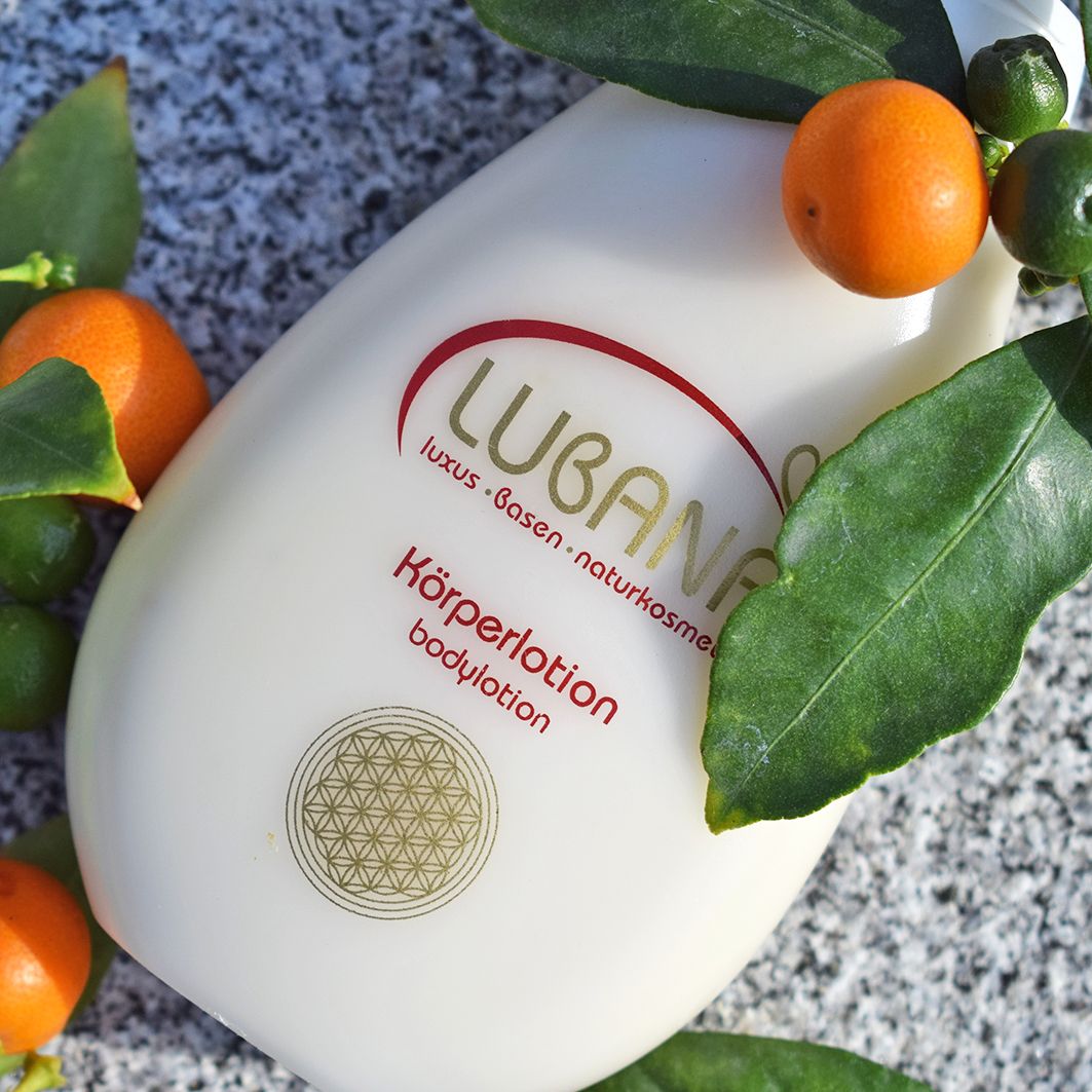 Beigefarbene Körperlotion-Flasche mit rotem Schriftzug Lubana. Umgeben von Blättern und kleinen Orangen. Goldener Kreis unten.