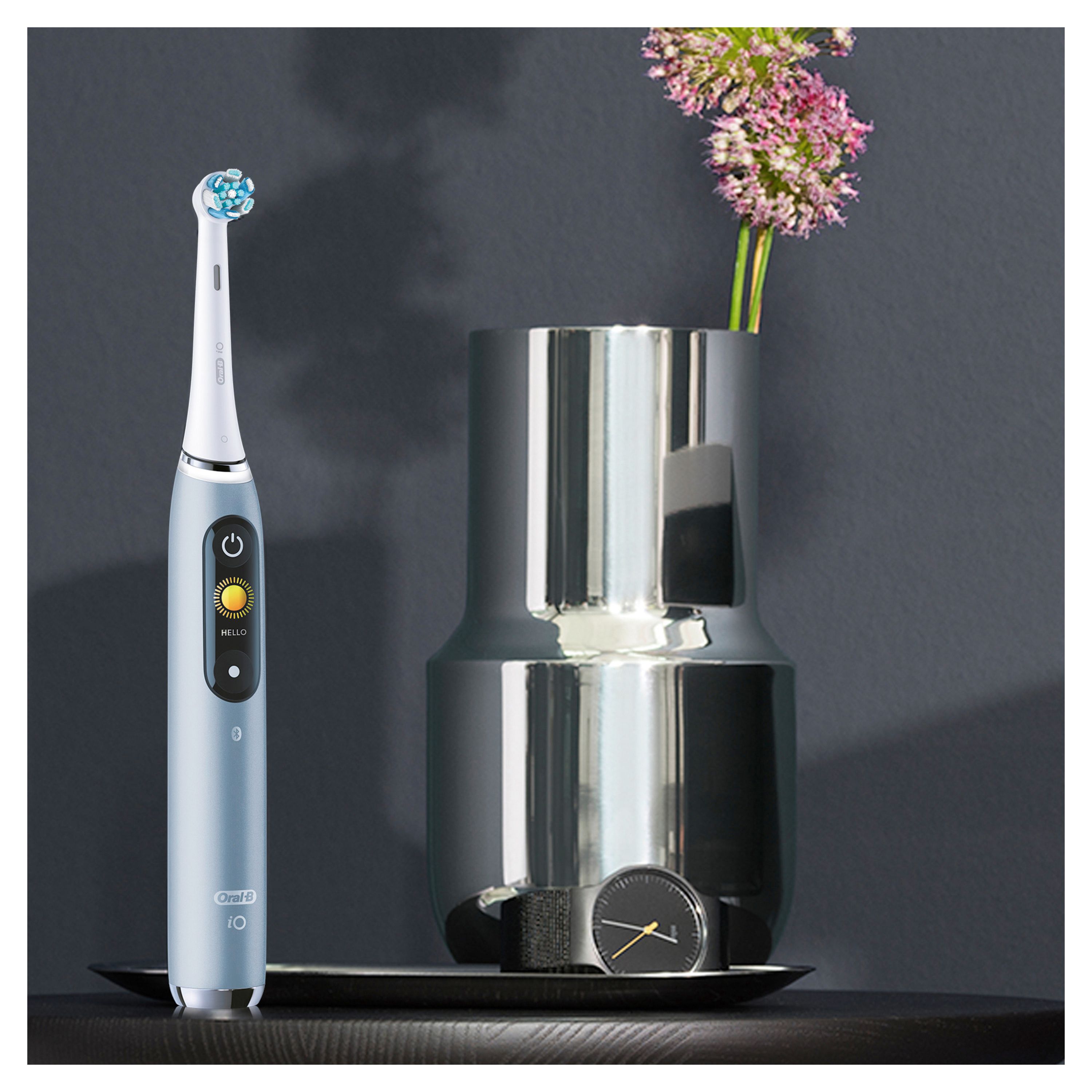 Blaue elektrische Zahnbürste neben Vase mit Blumen. Schwarzer Untergrund. Produktname und Marke nicht sichtbar.