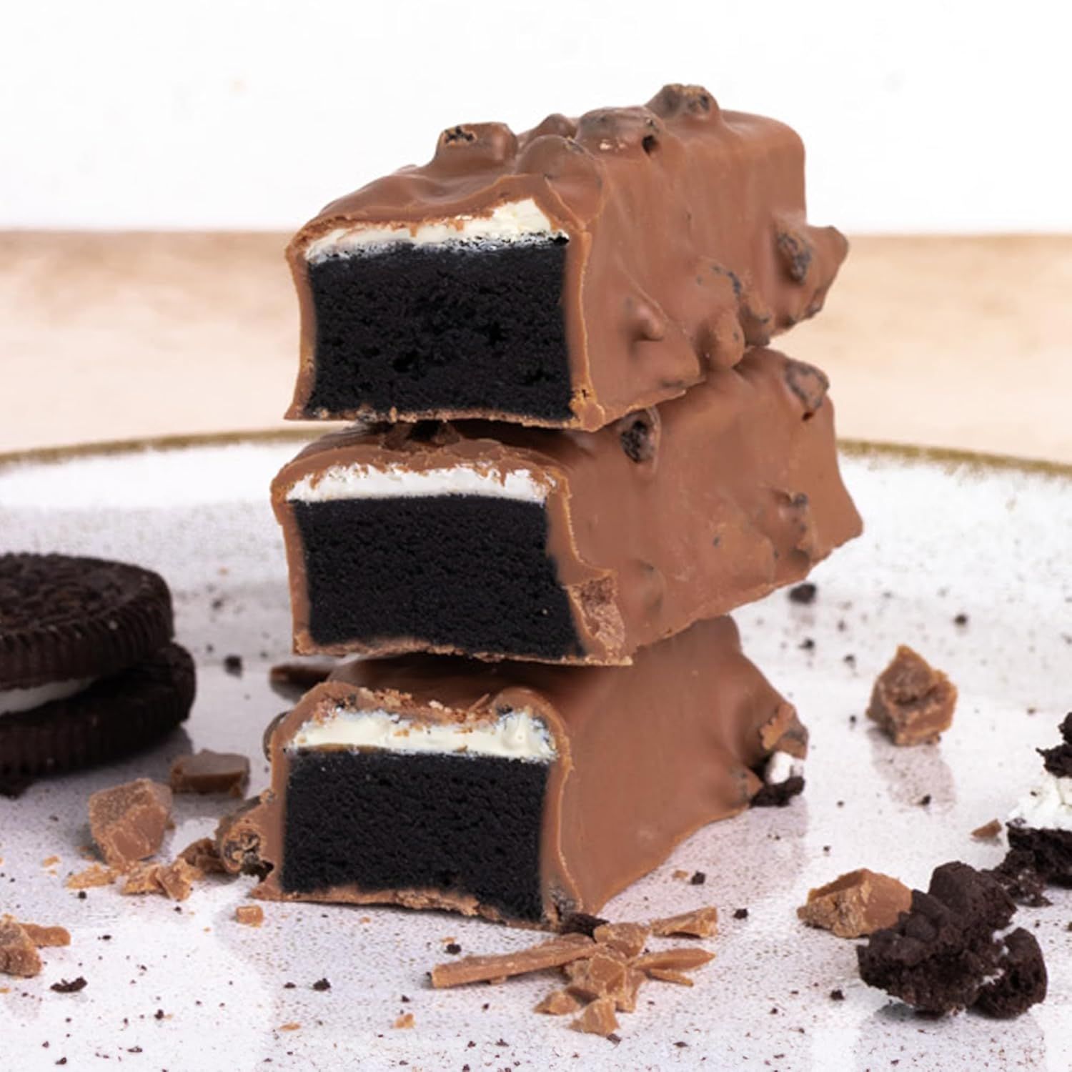 Drei angeschnittene Proteinriegel, Schokoladenüberzug mit Cookie-Stückchen. Innen dunkler Keks und helle Creme. Auf einem Teller.