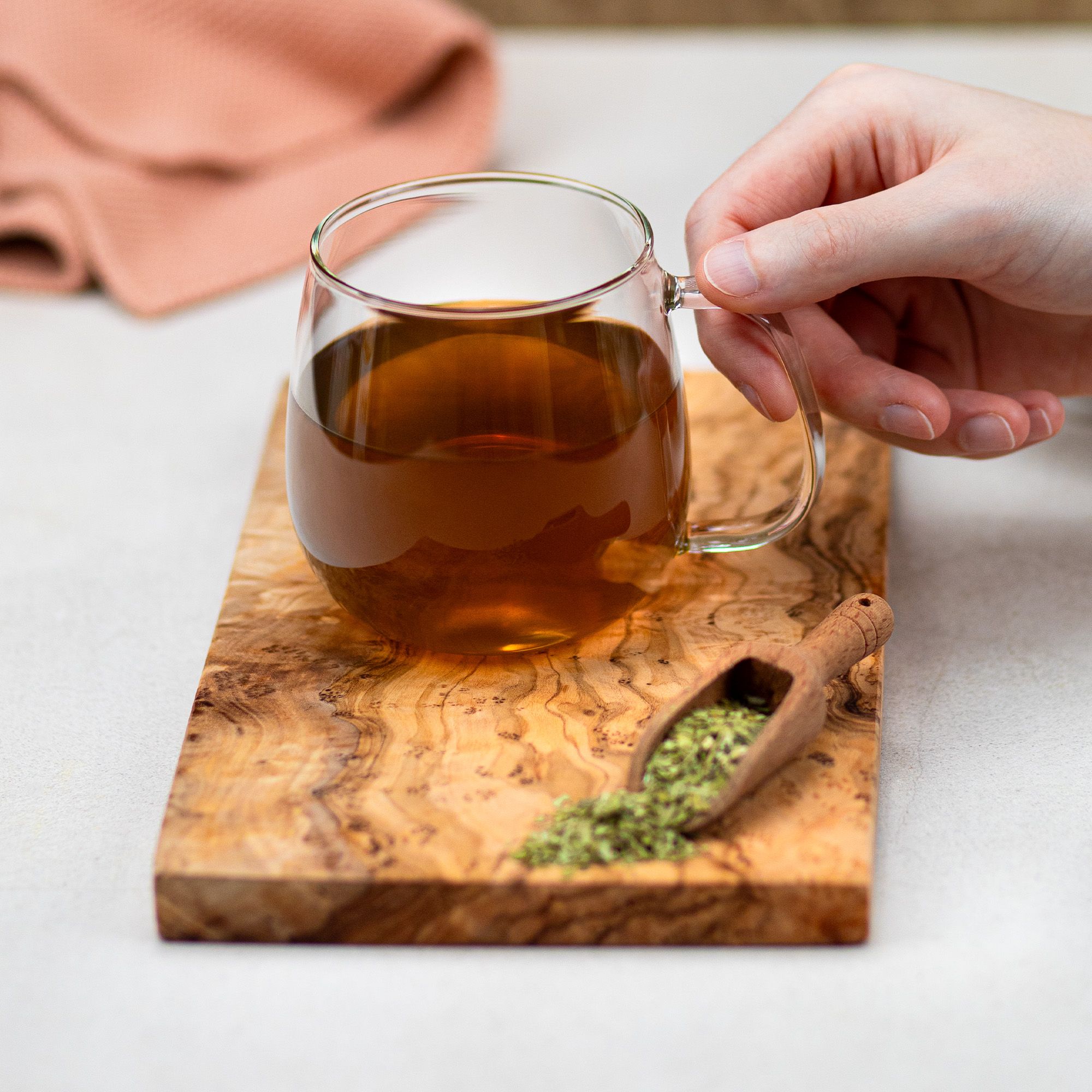 Glasbecher mit braunem Tee auf Holzbrett. Holzlöffel mit Mate-Tee daneben. Rosa Textil im Hintergrund.