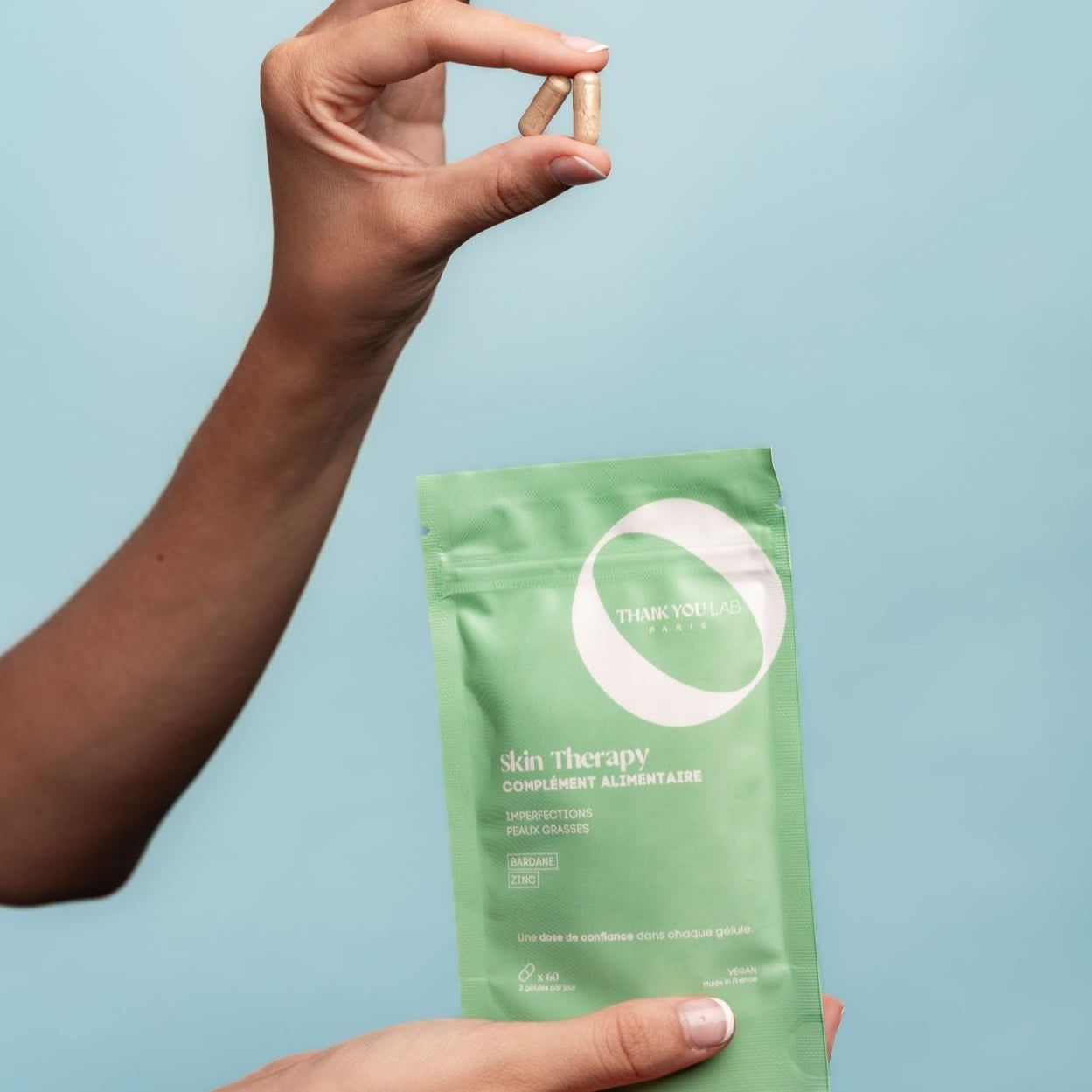 Grüne Verpackung mit Produktnamen und Kapseln. Eine Hand hält zwei Kapseln, die andere die Verpackung. Text: Skin Therapy, Complément Alimentaire.