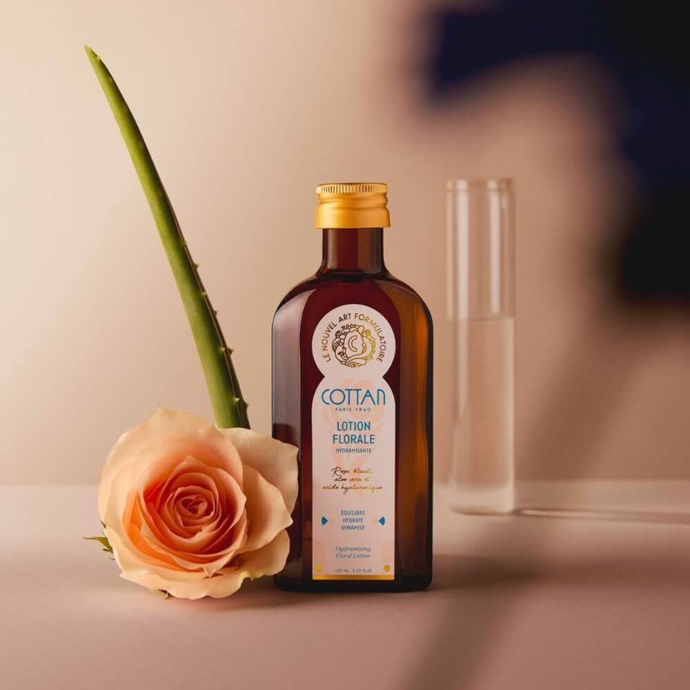 Braune Glasflasche mit goldenem Deckel und "COTTAN"-Logo. Eine Rose und ein Aloe-Blatt sind neben der Flasche platziert.