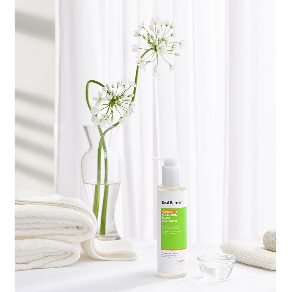 Flasche Real Barrier Control-T Cleansing Foam neben Handtüchern und Blumen. Auf weißem Hintergrund.