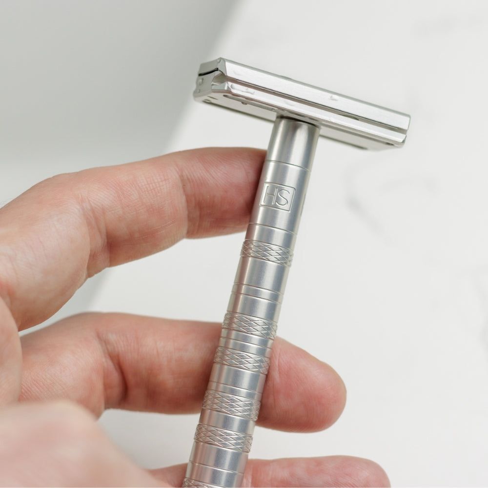 Sicherheitsrasierer AL13 Silber, Metall, in Hand gehalten. Griff mit HS-Logo und Rillen. Rasierkopf mit Klinge.