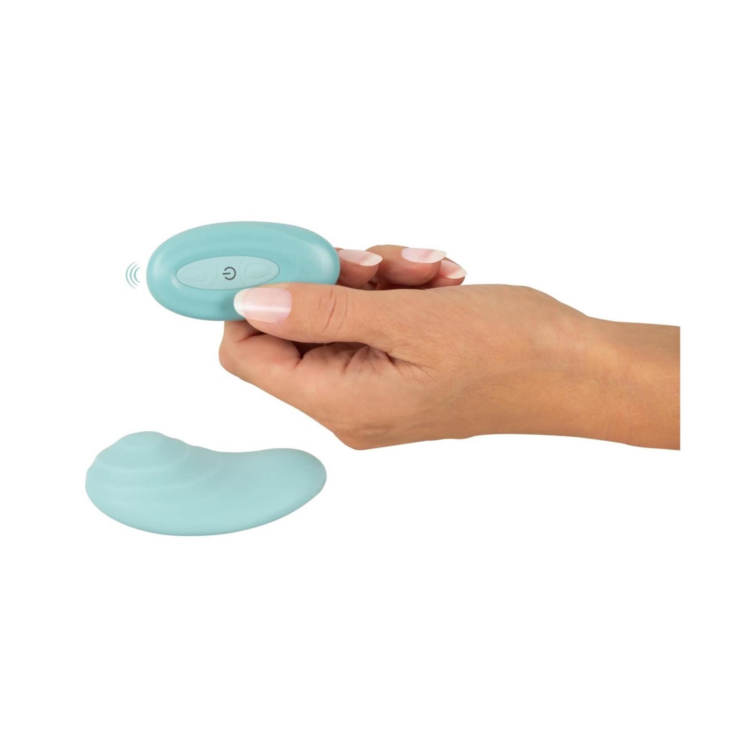 Hand hält hellblaue Fernbedienung. Vibrationsgerät liegt daneben. Funk-Symbol auf der Fernbedienung.