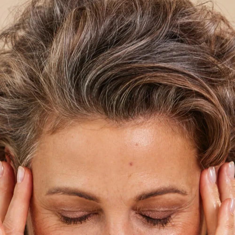 Frau mit geschlossenen Augen, Hände an den Schläfen. Graue Haare, braune Strähnen.