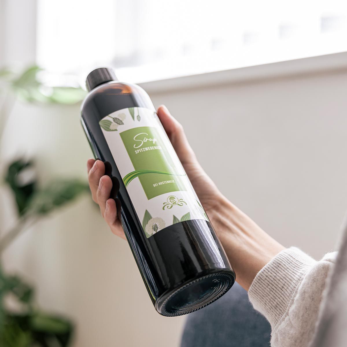 Person hält Flasche. Braune Flasche mit Etikett. Etikett mit grüner Schrift und floralen Elementen. Text: Sirup Spitzwegerich.