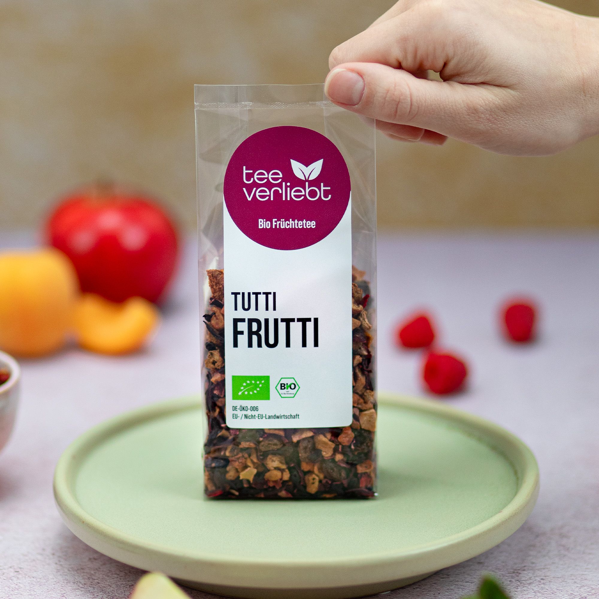 Verpackung mit losem Früchtetee auf einem Teller. Logo 'tee verliebt' und Text 'TUTTI FRUTTI'. Hand greift nach der Verpackung. Früchte im Hintergrund.