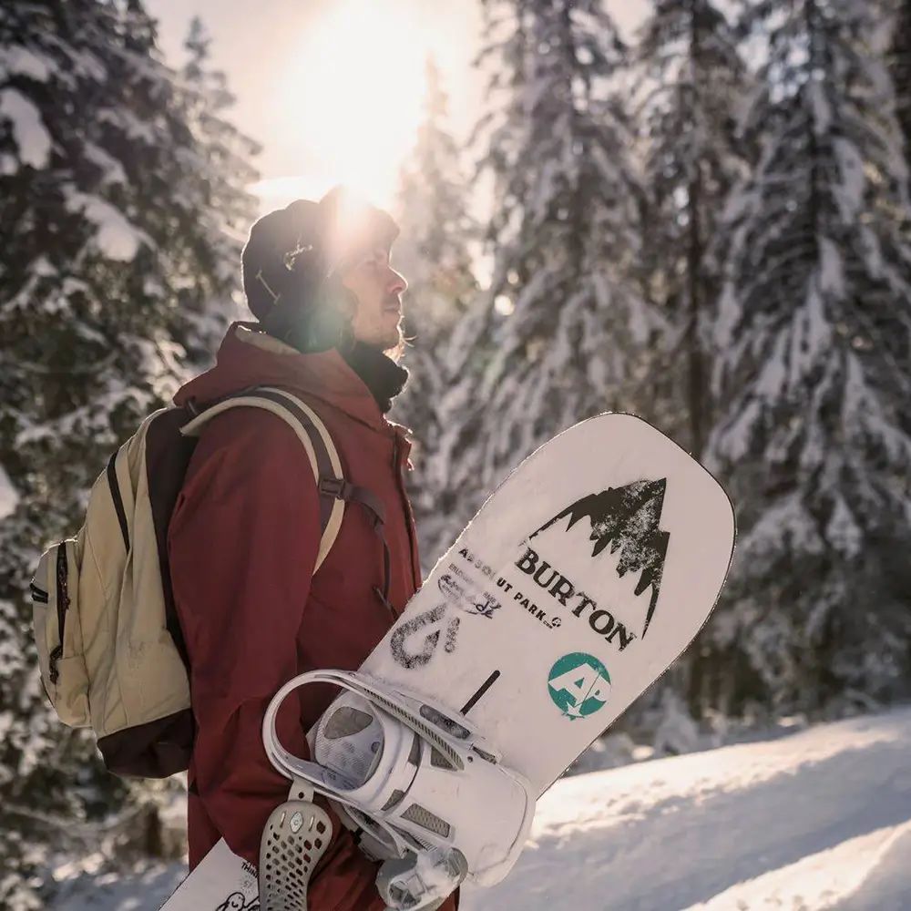 Person mit Rucksack und Snowboard in verschneiter Landschaft. Snowboard mit Aufschrift BURTON.