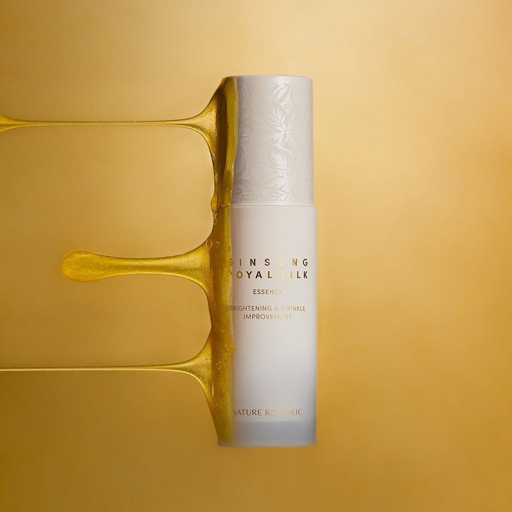 Beige Flasche mit Schriftzug GINSENG ROYAL SILK ESSENCE. Goldfarbene Flüssigkeit fließt über die Flasche. Marke: NATURE REPUBLIC.