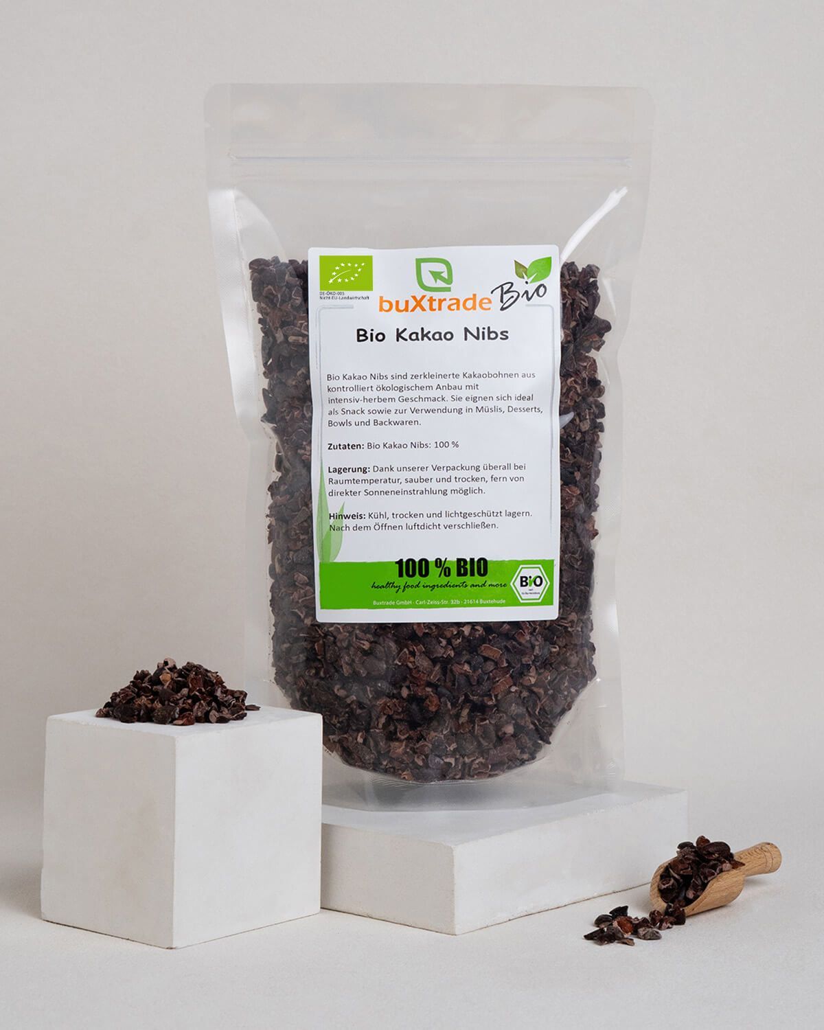 Bio Kakaonibs in transparenter Verpackung, daneben ein Holz-Löffel und ein Würfel mit Kakaonibs.