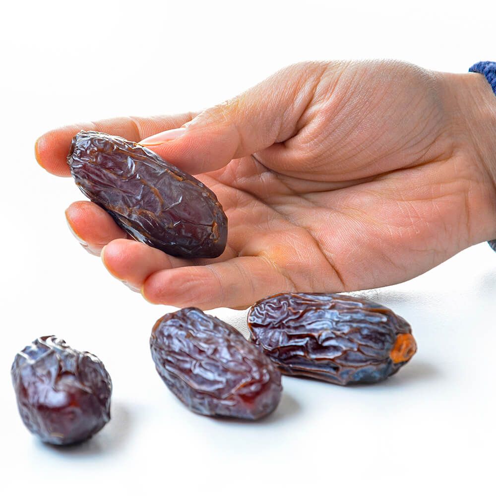 Hand hält eine Medjool-Dattel. Drei weitere Datteln liegen daneben. Helle Oberfläche.