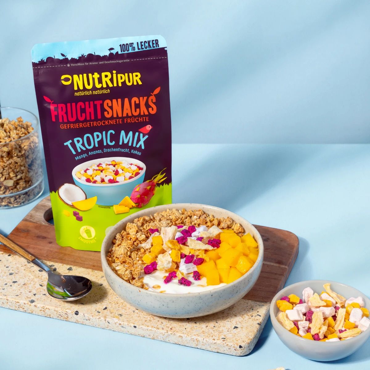 Beutel und Schalen mit gefriergetrockneten Früchten und Müsli. Aufschrift: NutriPur, Fruchtsnacks Tropic Mix. Auf einem Holzbrett.