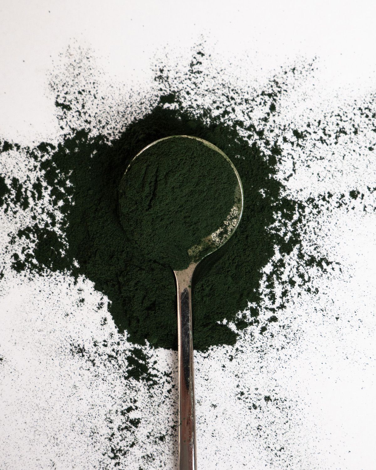 Ein Löffel mit Spirulina-Pulver auf weißem Hintergrund. Das Pulver ist um den Löffel herum verstreut.