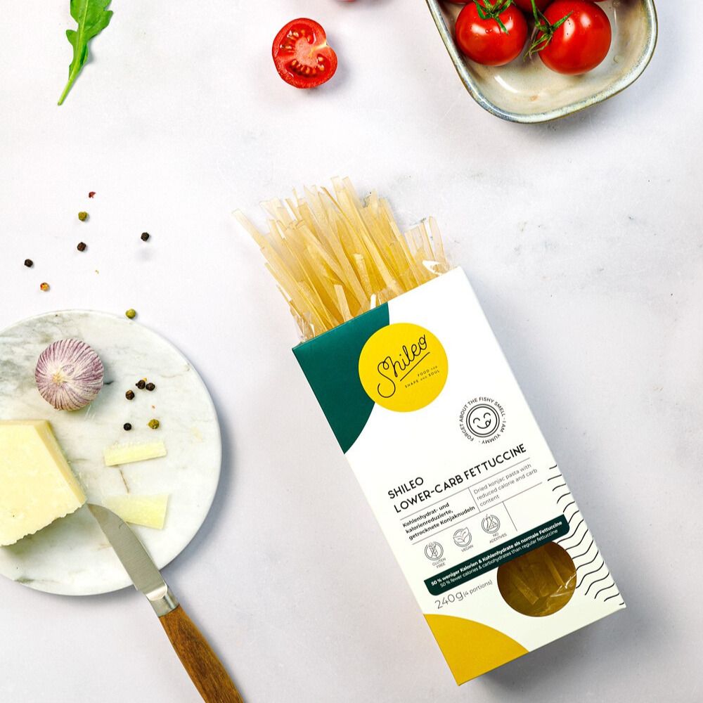 Verpackung SHILEO Lower-Carb Fettuccine neben Zutaten. Aufdrucke: Logo, Produktname, 240g. Auf dem Tisch: Tomaten, Käse, Knoblauch.