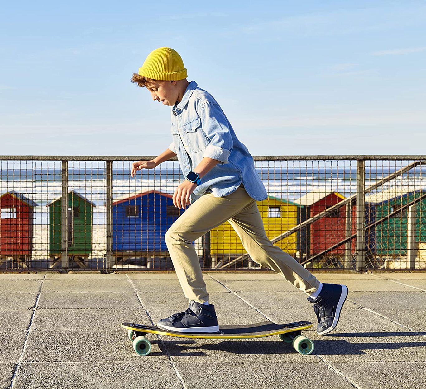 Junge auf einem Skateboard. Er trägt eine blaue Jeansjacke, eine gelbe Mütze und eine Smartwatch am Handgelenk.