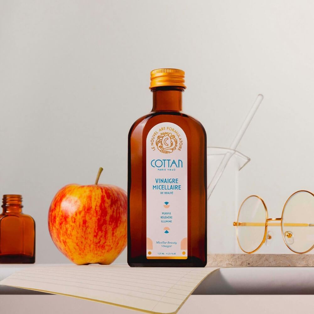 Braune Glasflasche mit goldfarbenem Verschluss. Weißes Etikett mit blauer Schrift. Neben Apfel, Becherglas und Brille.