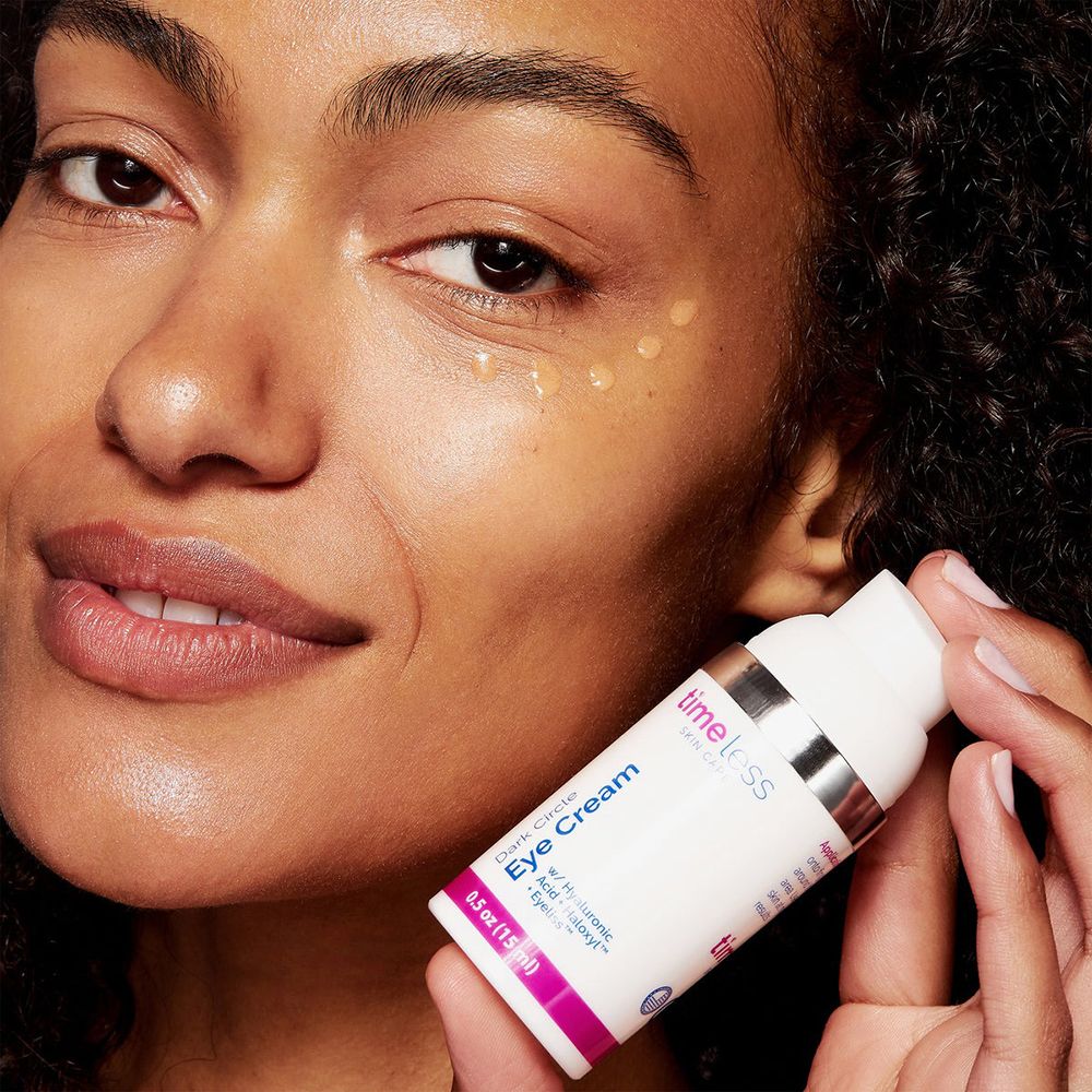 Frau hält eine Flasche. Aufschrift: TIMELESS SKIN CARE, Dark Circle Eye Cream, 0.5 oz (15 ml). Gel auf der Haut.