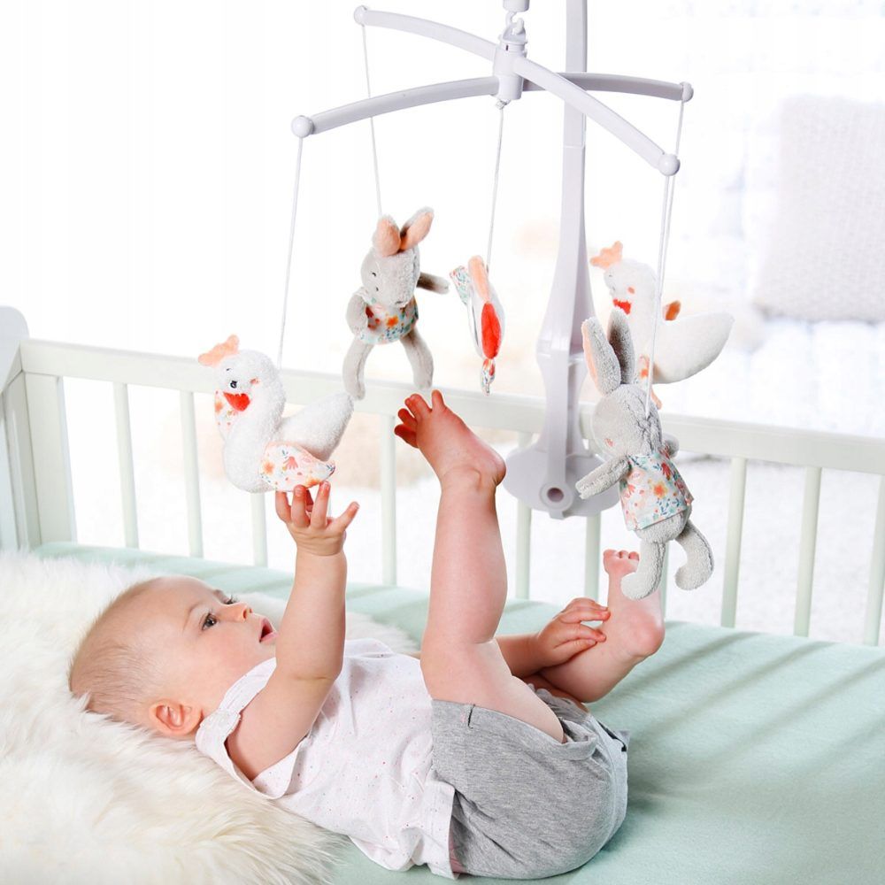 Baby liegt unter Mobile und greift danach. Mobile mit Hasen, Schwänen und Spiegel. Weißes Gestell. Rosa und blaue Akzente.
