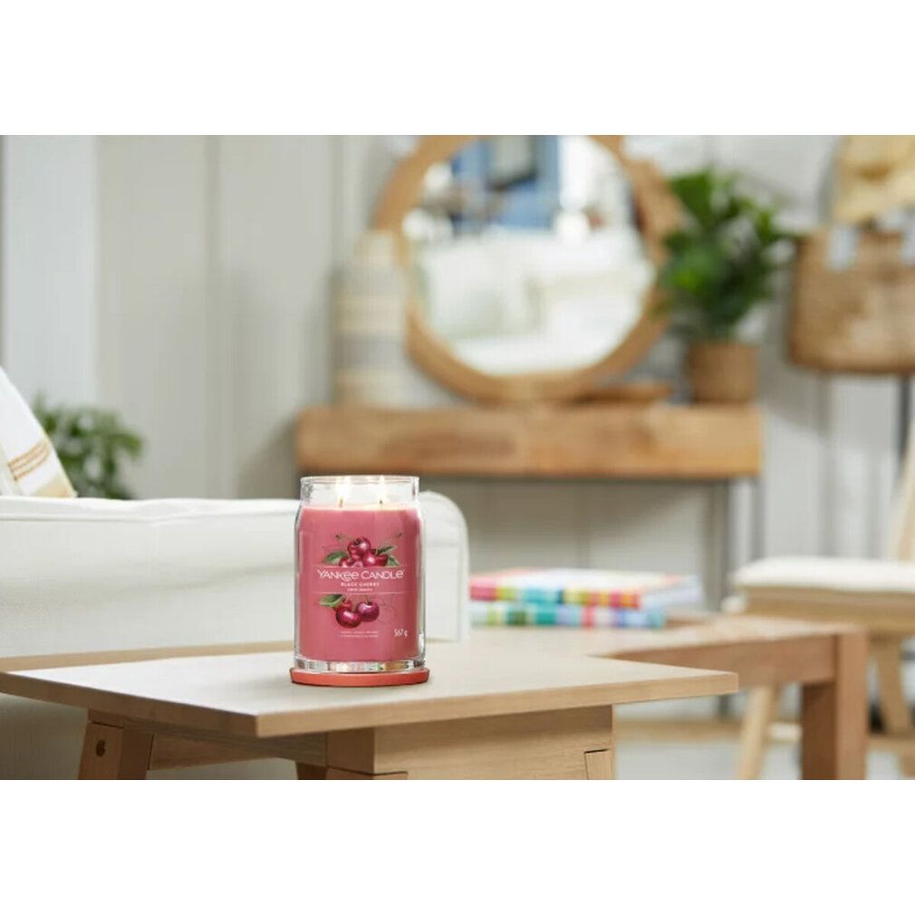 Kerze auf einem Tisch, im Hintergrund unscharfe Wohnzimmereinrichtung. 'Yankee Candle Black Cherry' mit Kirschabbildung.
