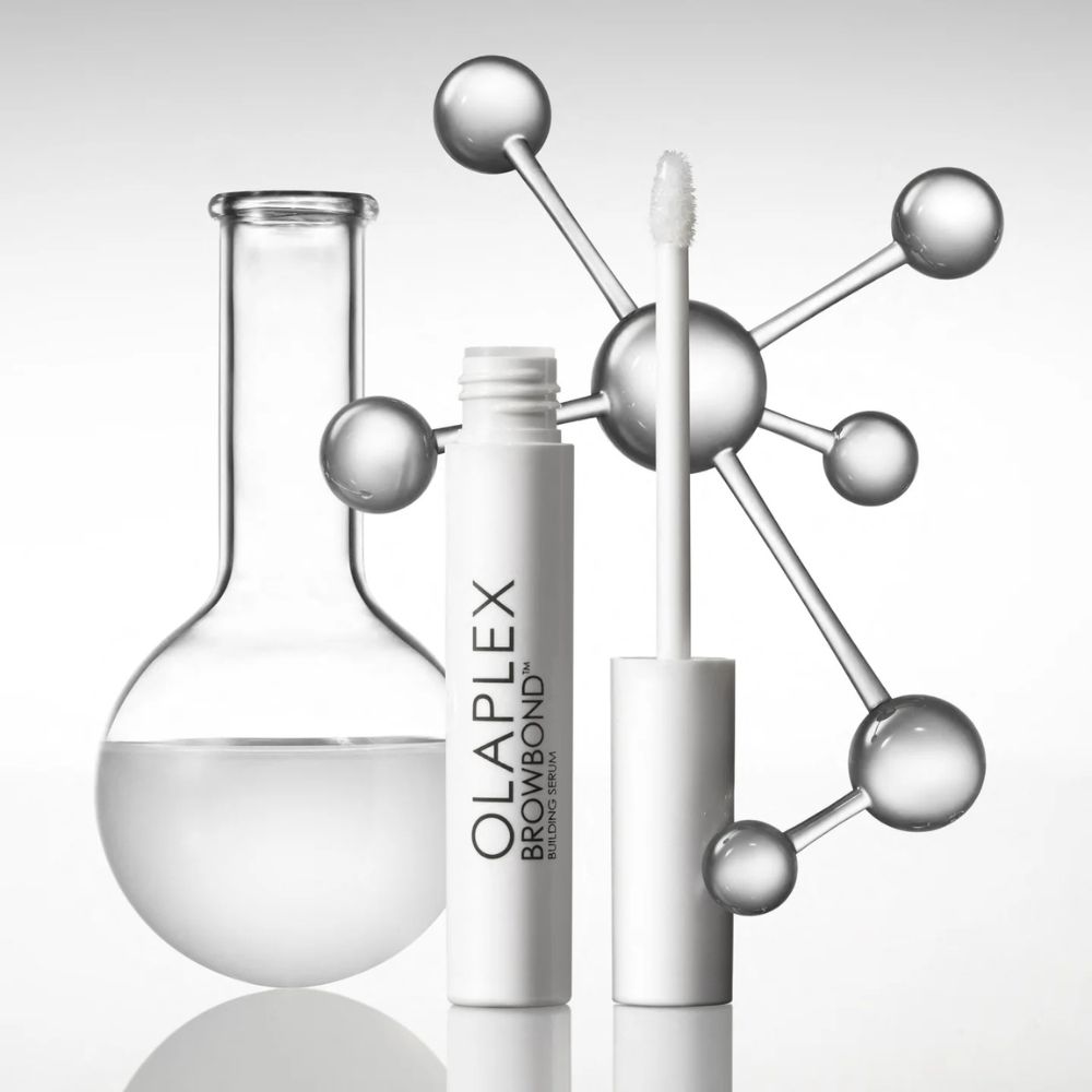 Weißes Serumfläschchen mit Applikator. OLAPLEX BROWBOND BUILDING SERUM. Neben einem Reagenzglas und einer Molekülstruktur.