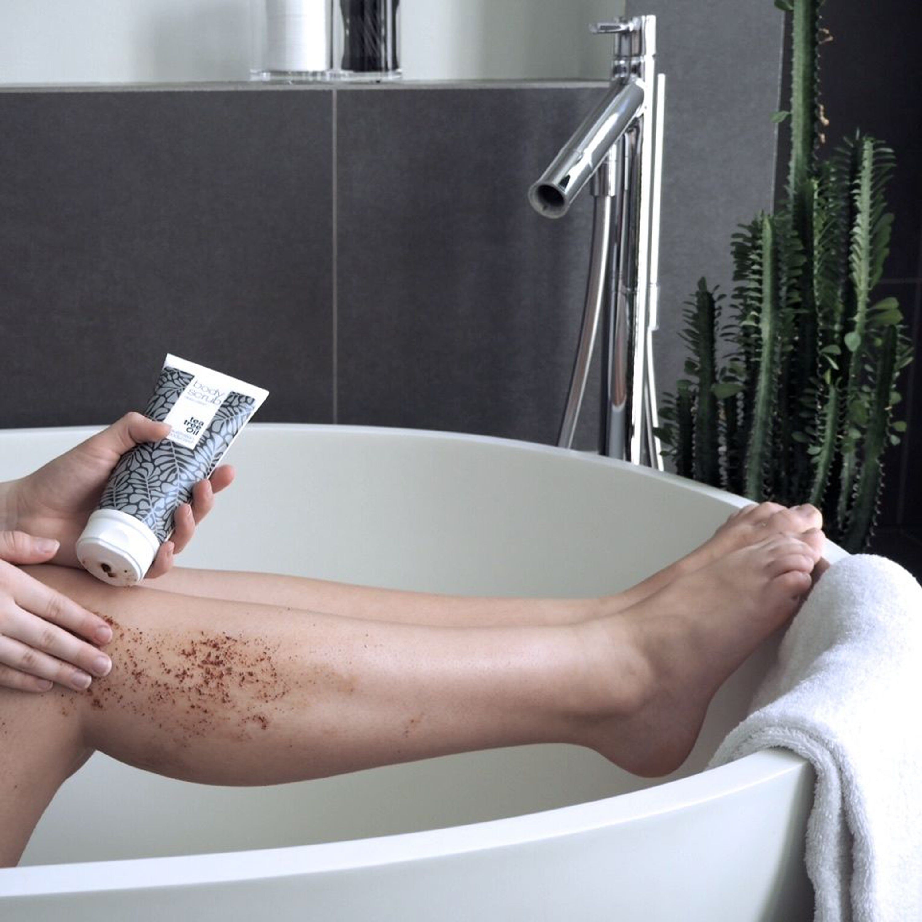 Person hält Tube „Body Scrub“ über Bein in Badewanne. Peeling auf Bein. Marke: Australian Bodycare.