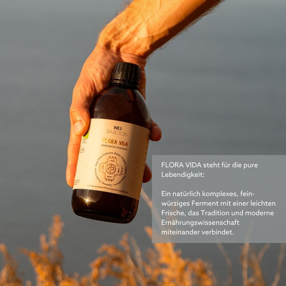 Braune Glasflasche mit Etikett, gehalten von einer Hand. Aufschrift: FLORA VIDA. Hintergrund: Wasser und Pflanzen.