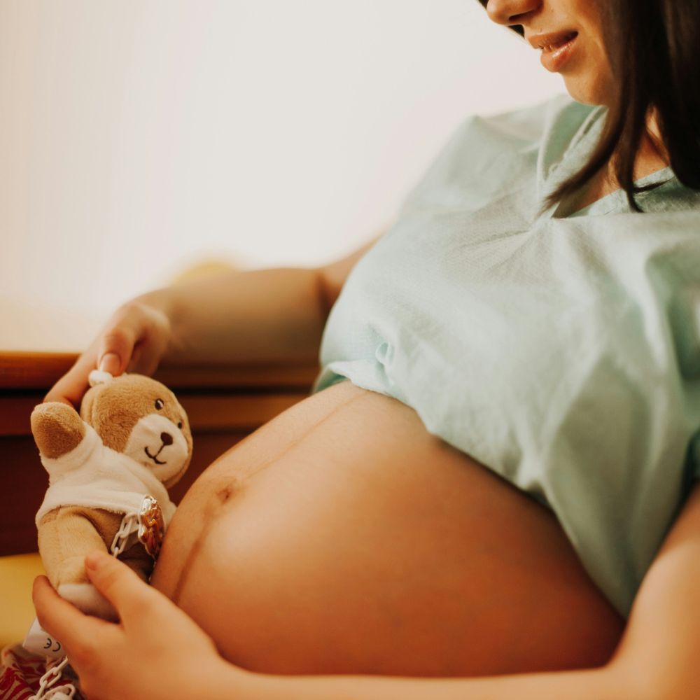 Schwangere Frau mit Teddybär auf dem Bauch. Sie trägt ein hellgrünes Oberteil.