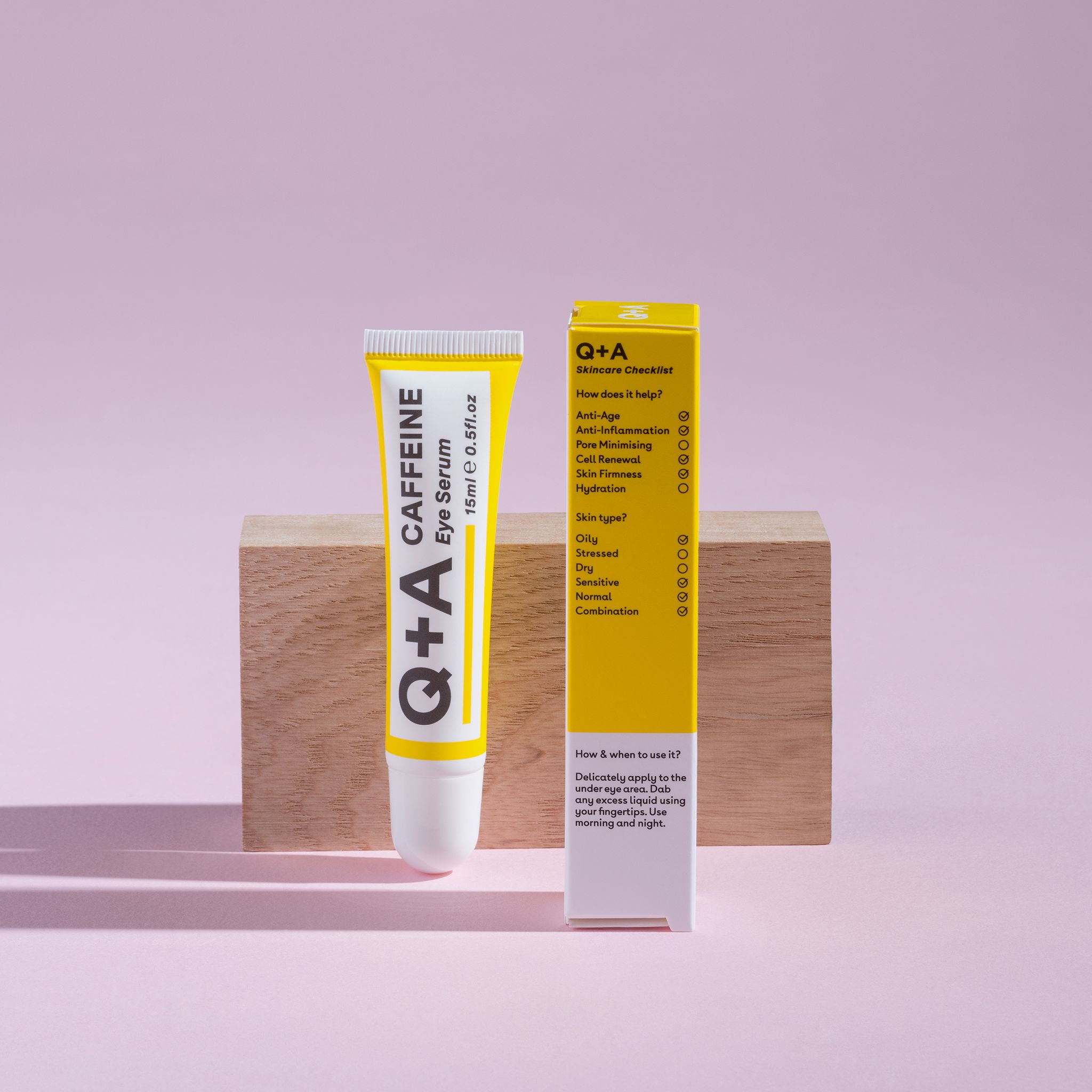 Gelbe Tube und Schachtel. Aufschrift: Q+A, CAFFEINE Eye Serum, 15ml 0.5fl.oz. Auf rosa Untergrund mit Holzblock.