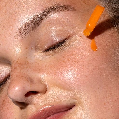 Gesicht einer Frau mit geschlossenen Augen. Ein Tropfen orangefarbenes Serum wird mit einer Pipette auf die Haut aufgetragen.