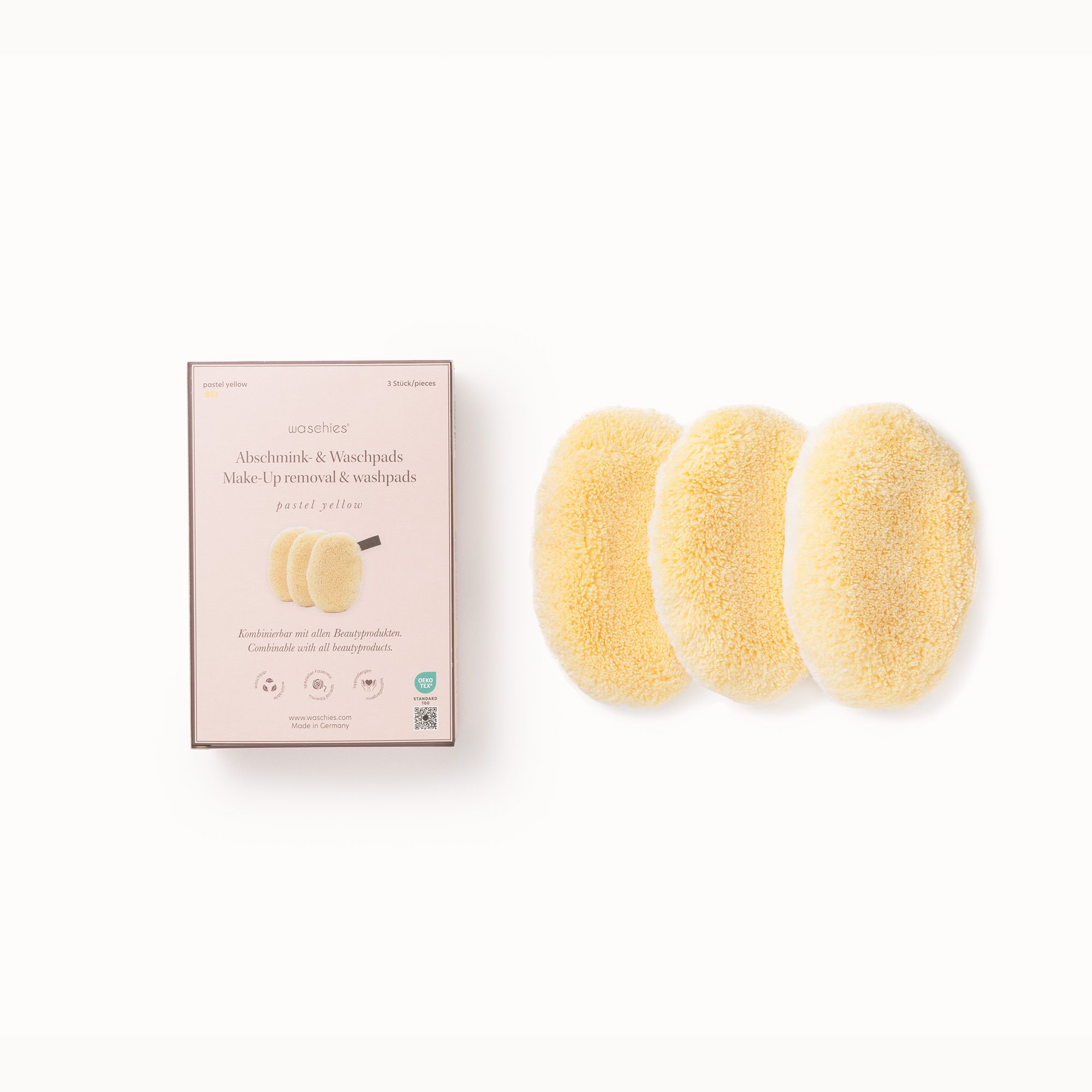 Drei gelbliche, ovale Abschminkpads neben einer Verpackung. Verpackung mit Produktname und Text. Pads aus Textilmaterial.