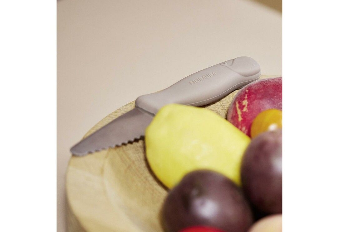 Beiges FILIBABBA Kindermesser mit wellenförmiger Klinge liegt neben Obst in einer Holzschale. Gelbe und rote Früchte.