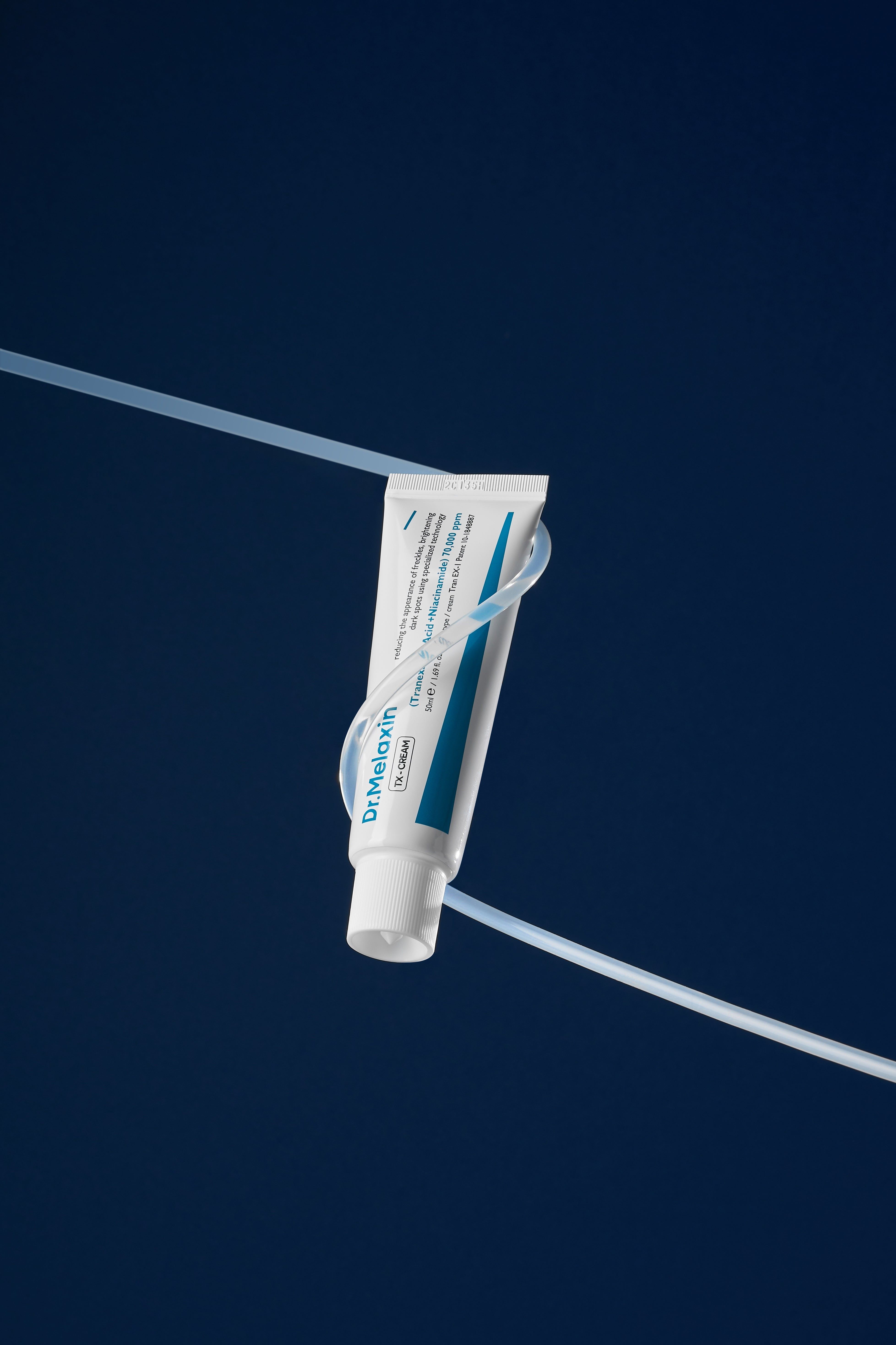 Weiße Tube mit blauer Beschriftung, umwickelt von einer durchsichtigen Schnur. Aufschrift: Dr.Melaxin TX-CREAM.