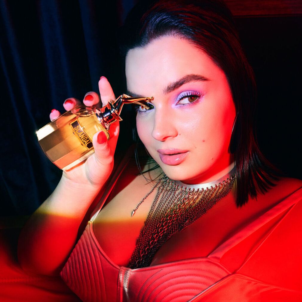 Frau hält Parfumflakon mit goldenem Verschluss. Flakon mit Schriftzug Scandal. Frau trägt rotes Oberteil und Schmuck.