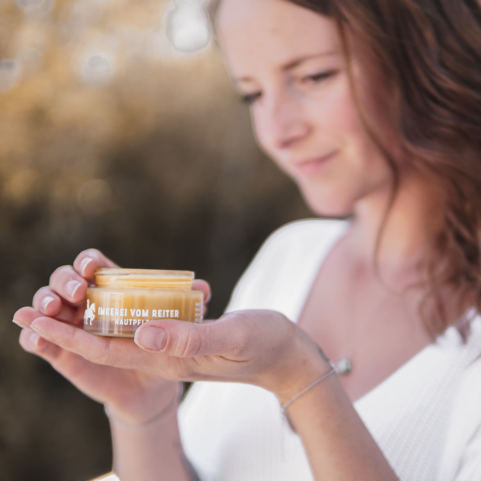 Frau hält Creme-Tiegel. Aufschrift: Imkerei vom Reiter Hautpflege. Gelbe Creme sichtbar. Logo: Reiter auf Pferd.