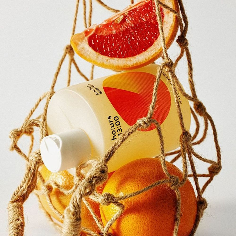 Flasche in einem Netzbeutel mit Früchten. Flasche: ho:urs, Sun Step Body Wash. Früchte: Orangen, Grapefruit.