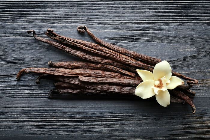 Dunkelbraune Vanilleschoten auf Holzuntergrund. Eine Vanilleblüte liegt daneben.