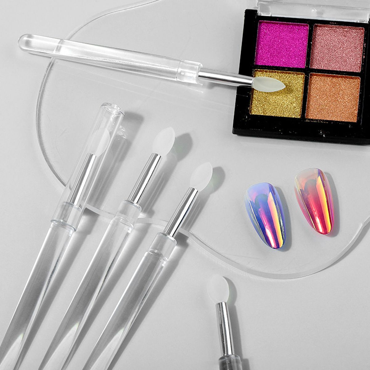 Mehrere transparente Applikatoren mit Silikonspitzen neben einem Lidschatten-Quad und holografischen Nägeln.