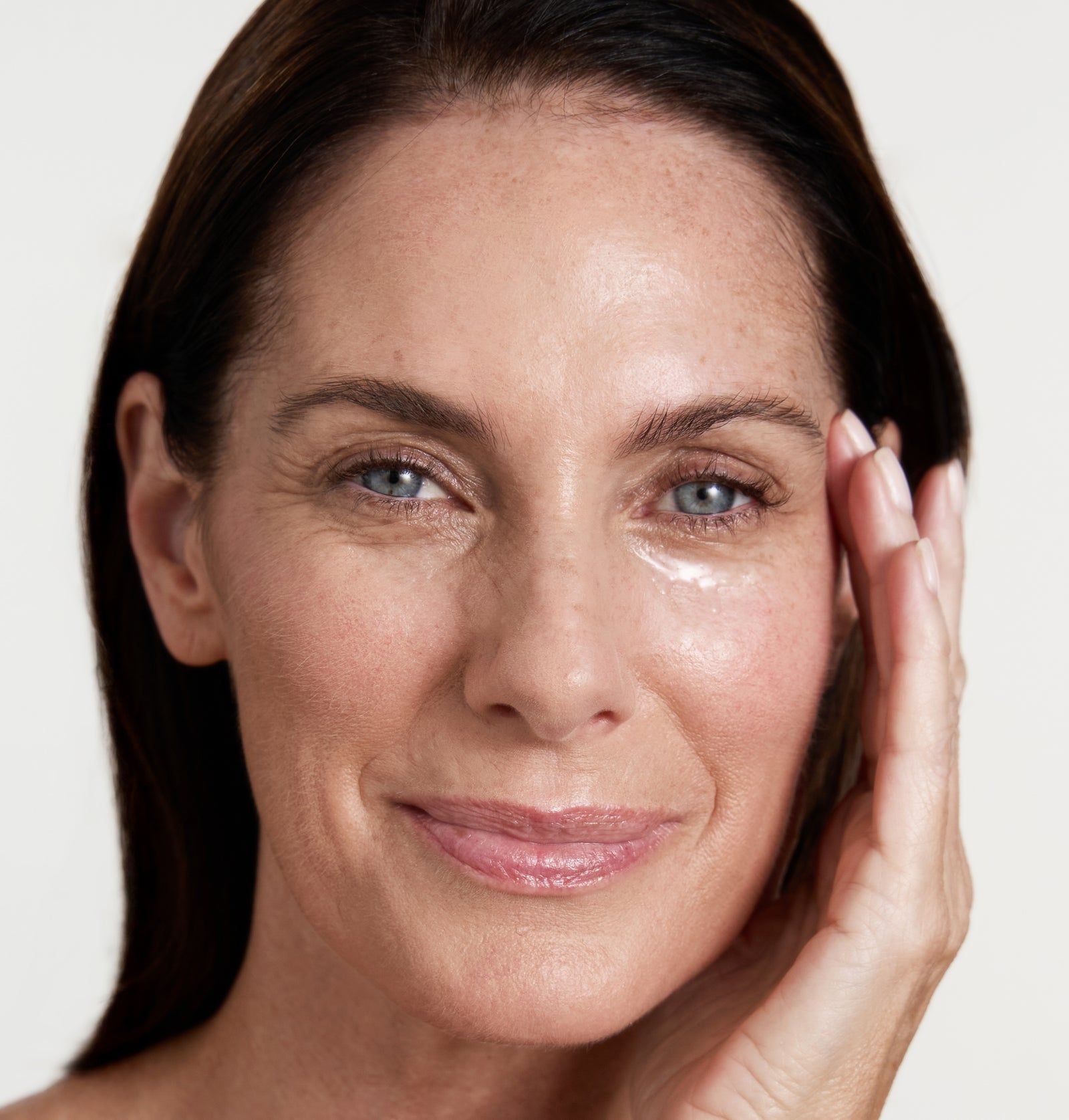 Frau mit Hautpflegeprodukt unter dem Auge. Hand berührt Gesicht. Helle Haut, braune Haare.