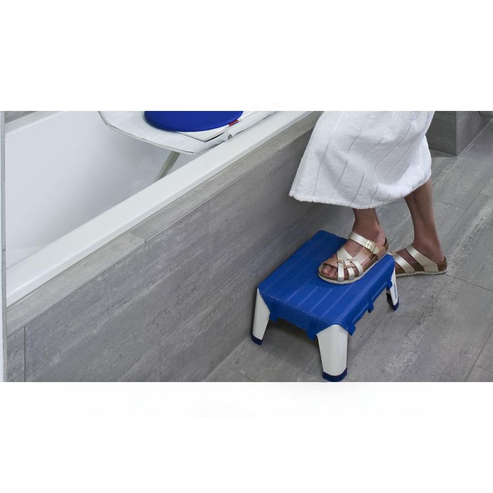 Person steigt mit Hilfe eines blauen Tritts in eine Badewanne. Weißes Gestell, rutschfeste Oberfläche.