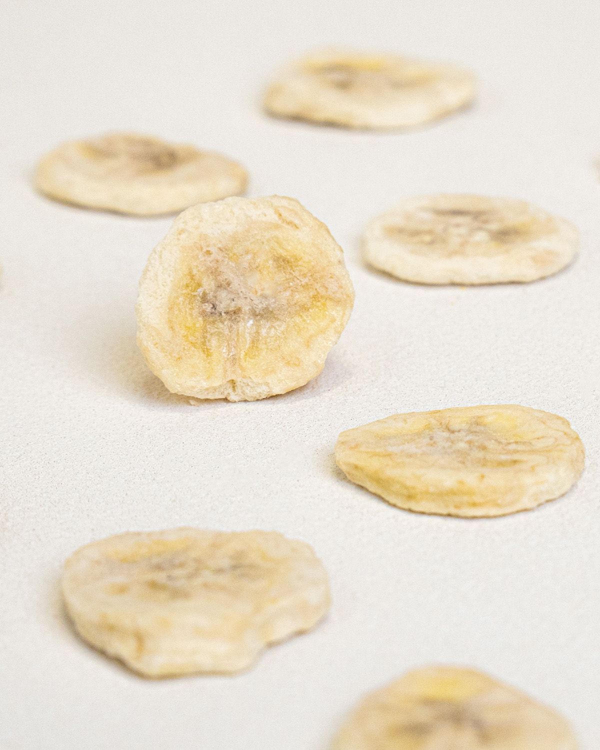 Nahaufnahme von gefriergetrockneten Bananenscheiben auf weißem Untergrund. Einzelne Scheiben, leicht gelblich.