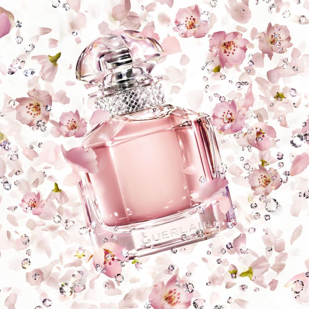 Parfümflakon mit rosa Flüssigkeit, umgeben von rosa Blütenblättern und Kristallen. Marke Guerlain.