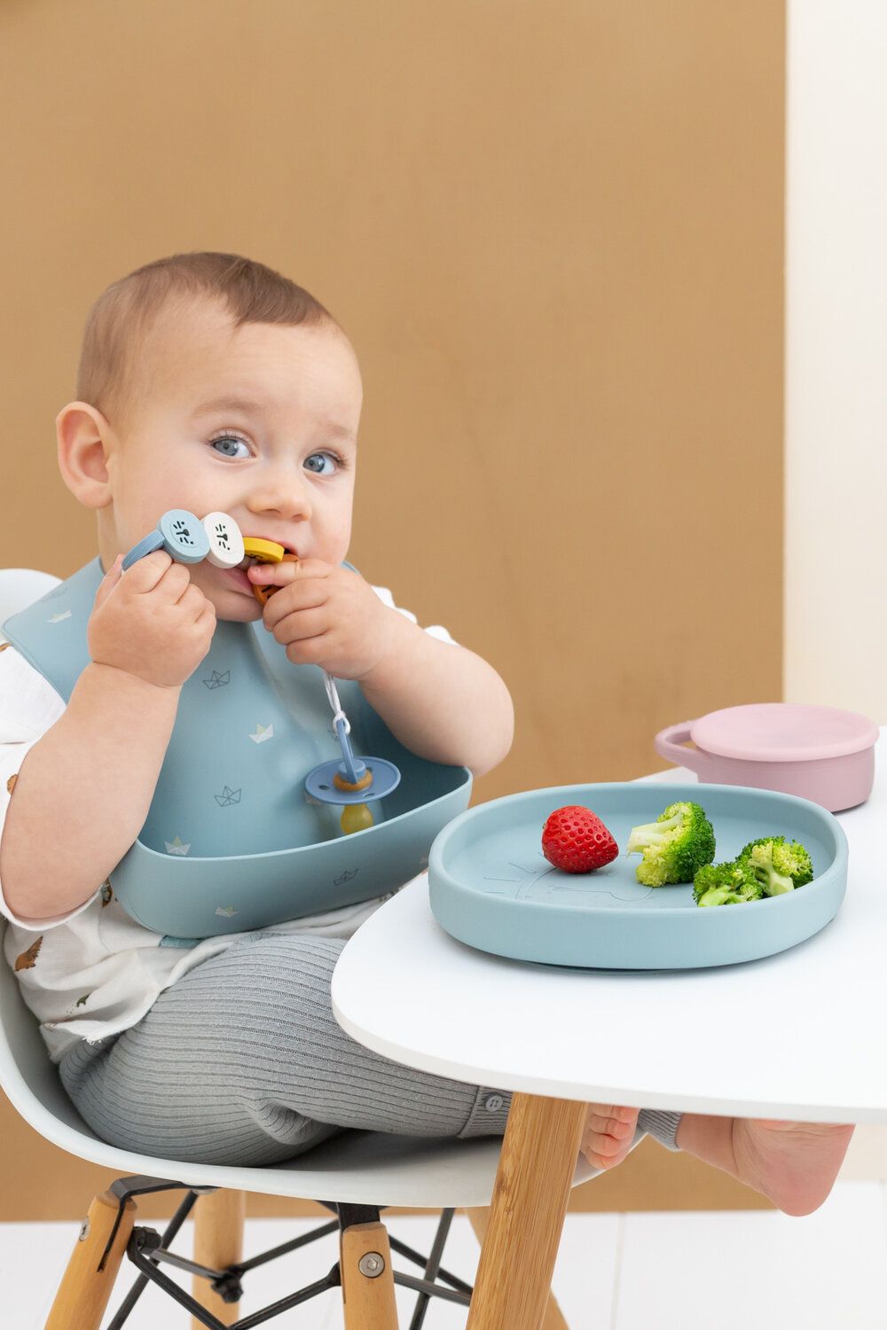 Baby am Tisch mit Teller, Lätzchen und Schnuller. Teller mit Erdbeere und Brokkoli. Lätzchen und Teller in Blau.
