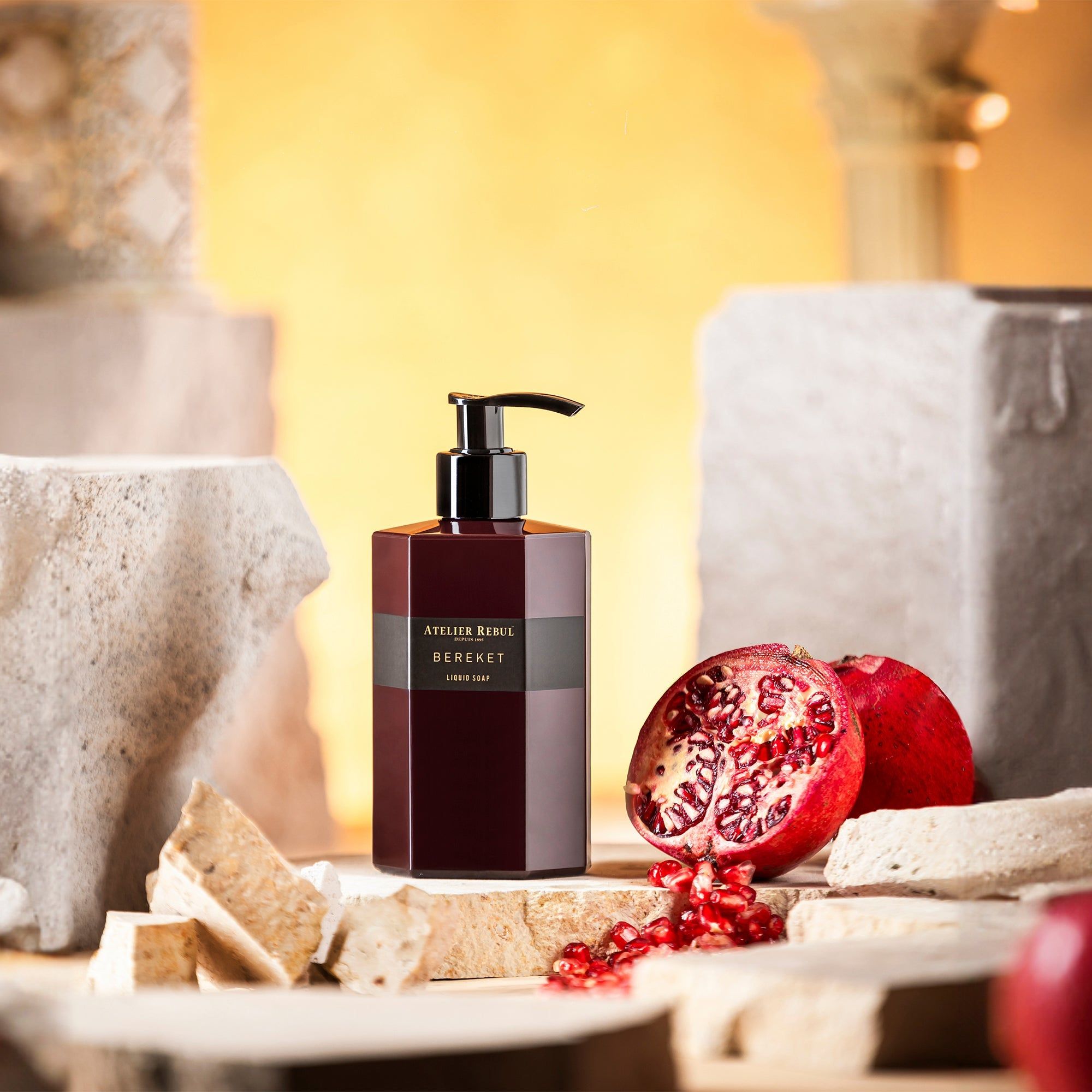 Bordeauxfarbene Flüssigseife mit schwarzem Pumpkopf. Granatapfel und Steinobjekte im Hintergrund. Aufschrift: Atelier Rebul Bereket.
