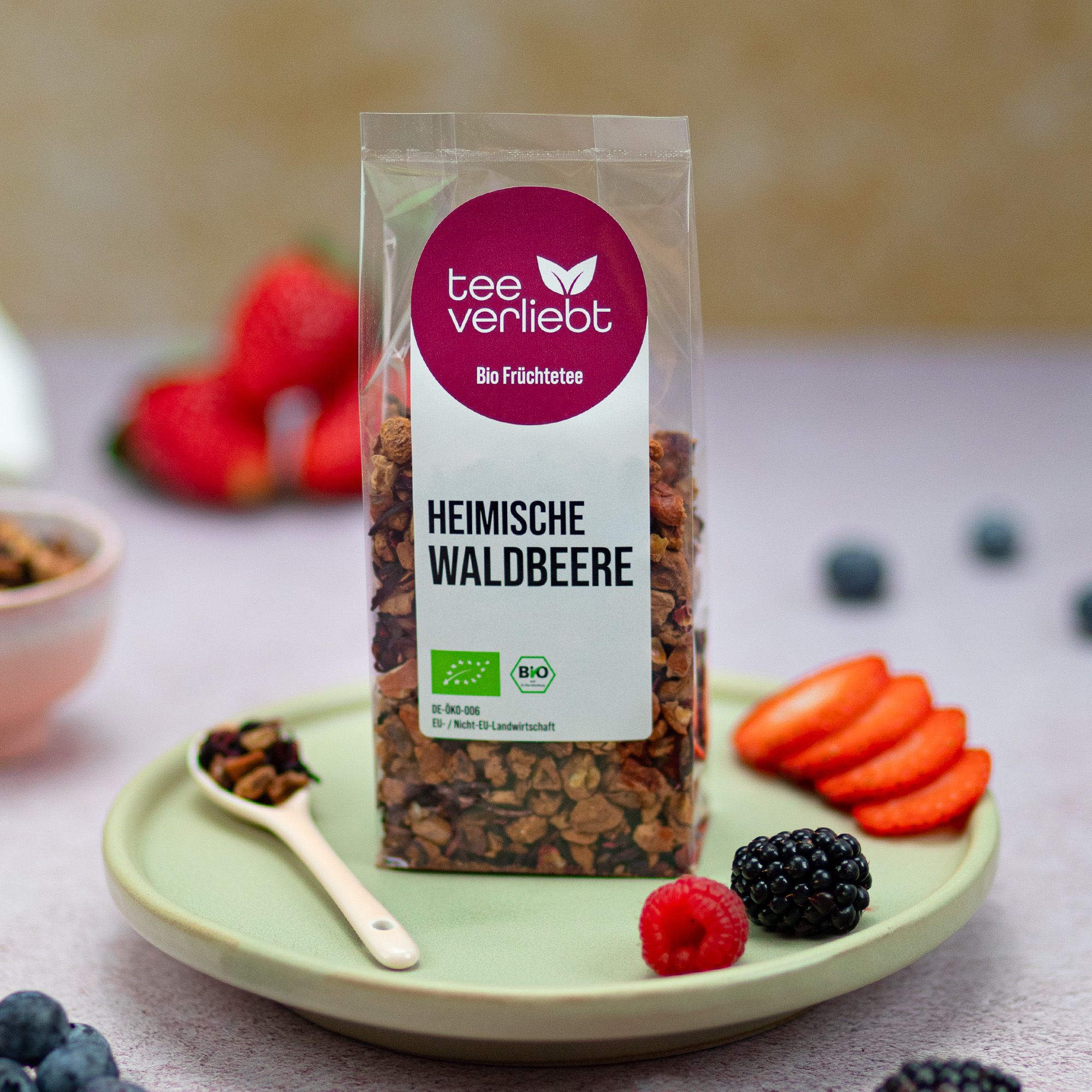 Verpackung mit losem Früchtetee, daneben Beeren und eine Schale mit Tee. Auf einem Teller liegt ein Löffel mit Tee.