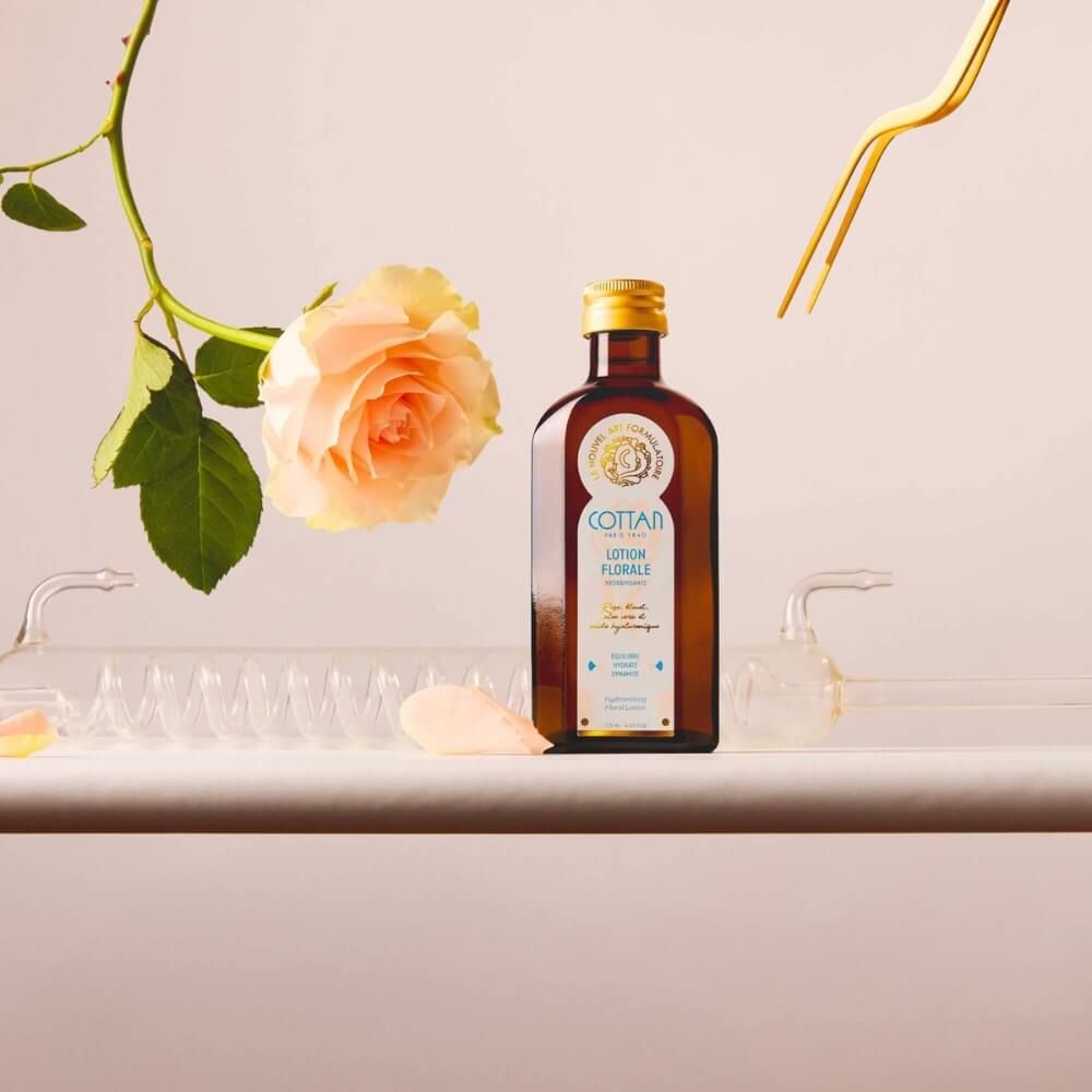 Braune Glasflasche mit goldenem Deckel und "COTTAN"-Logo. Eine Rose und Laborutensilien sind im Bild zu sehen.