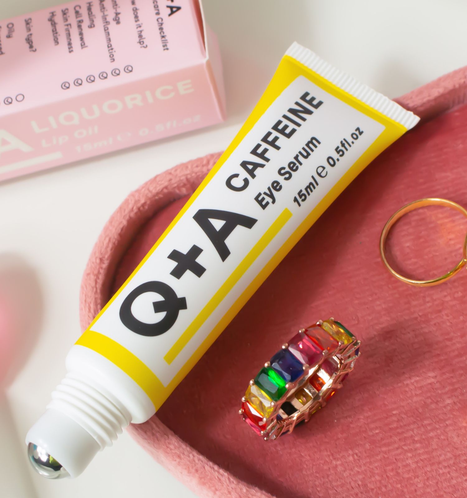 Gelbe Tube mit Text. Aufschrift: Q+A, CAFFEINE Eye Serum, 15ml 0.5fl.oz. Auf rosa Untergrund mit Schmuck.